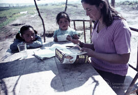 Mujer recortando una caja de cartón, junto a dos niñas pequeñas